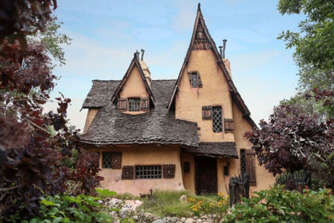 A storybook-style cottage with steep, curved shingled roofs and arched wooden windows sits among trees and flowering shrubs.