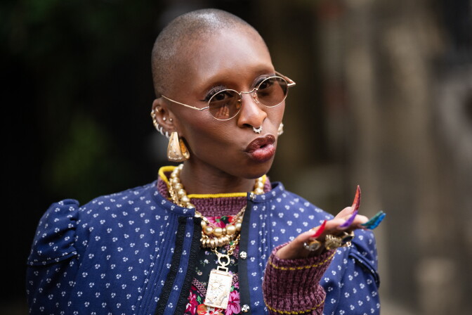 A bald person with round glasses, layered gold necklaces, and colorful nail art gestures while speaking outdoors.