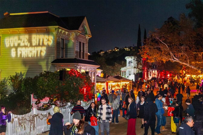A crowd walks a street lined with Victorian houses at night, one lit with the words 'Boo-ze, Bites & Frights' for a Halloween event.