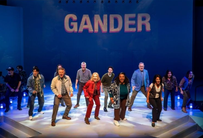 Performers dance on a stage in a pose with one leg raised, under a blue backdrop reading 'GANDER'.