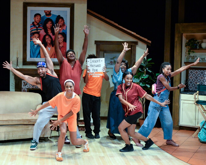 Actors in a stage play strike playful poses with arms raised in a sitcom-style living room set, one holding a sign reading 'Insert Hayward Here'.