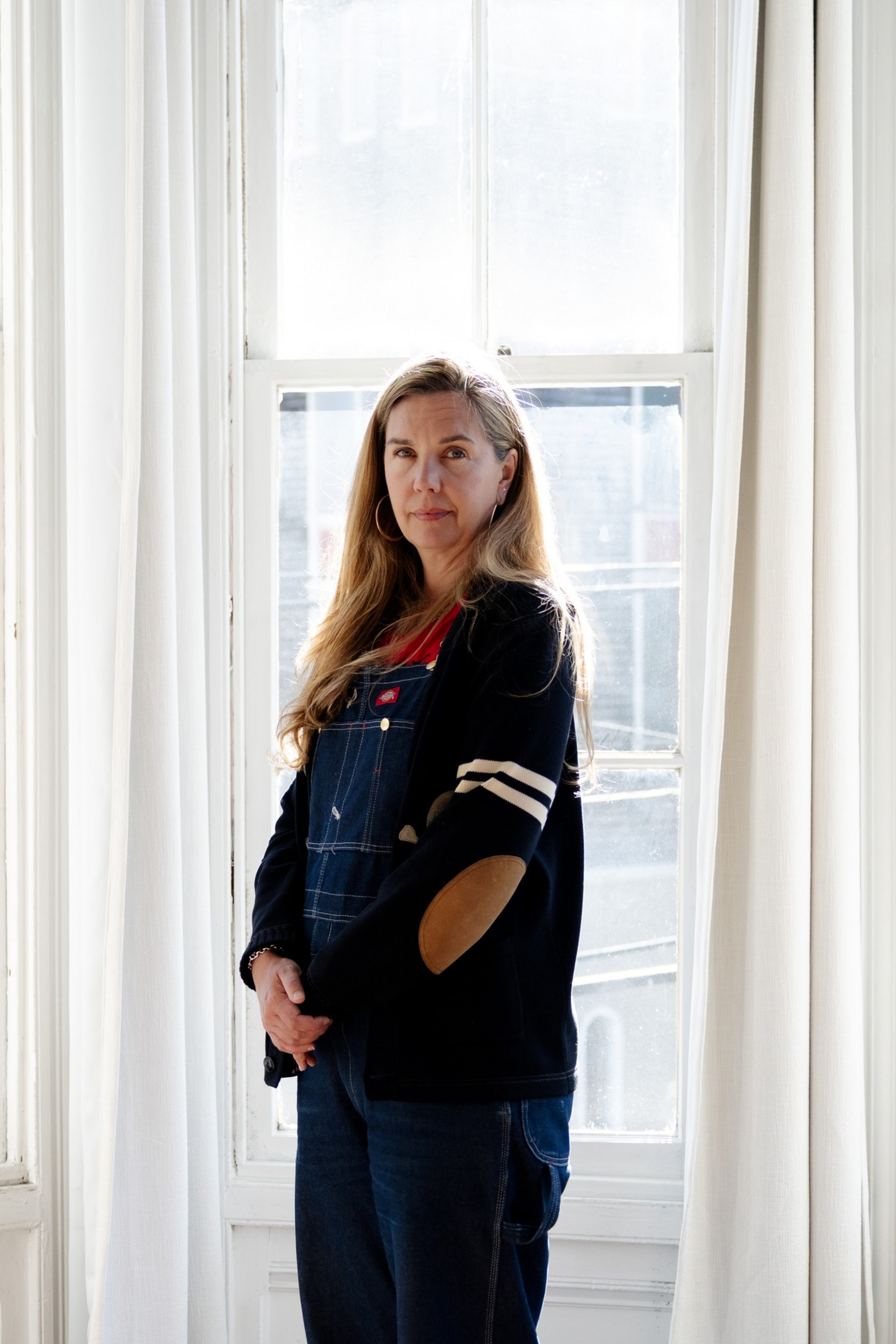 A woman with long blonde hair wears denim overalls and a black jacket with beige elbow patches, standing calmly in front of a large bright window.