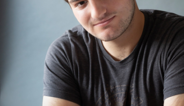 A young man with short brown hair and light facial hair leans forward with his arms crossed, wearing a dark gray T-shirt against a plain background.