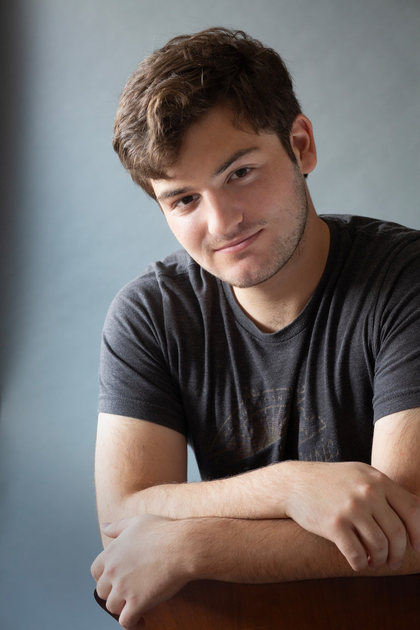 A young man with short brown hair and light facial hair leans forward with his arms crossed, wearing a dark gray T-shirt against a plain background.