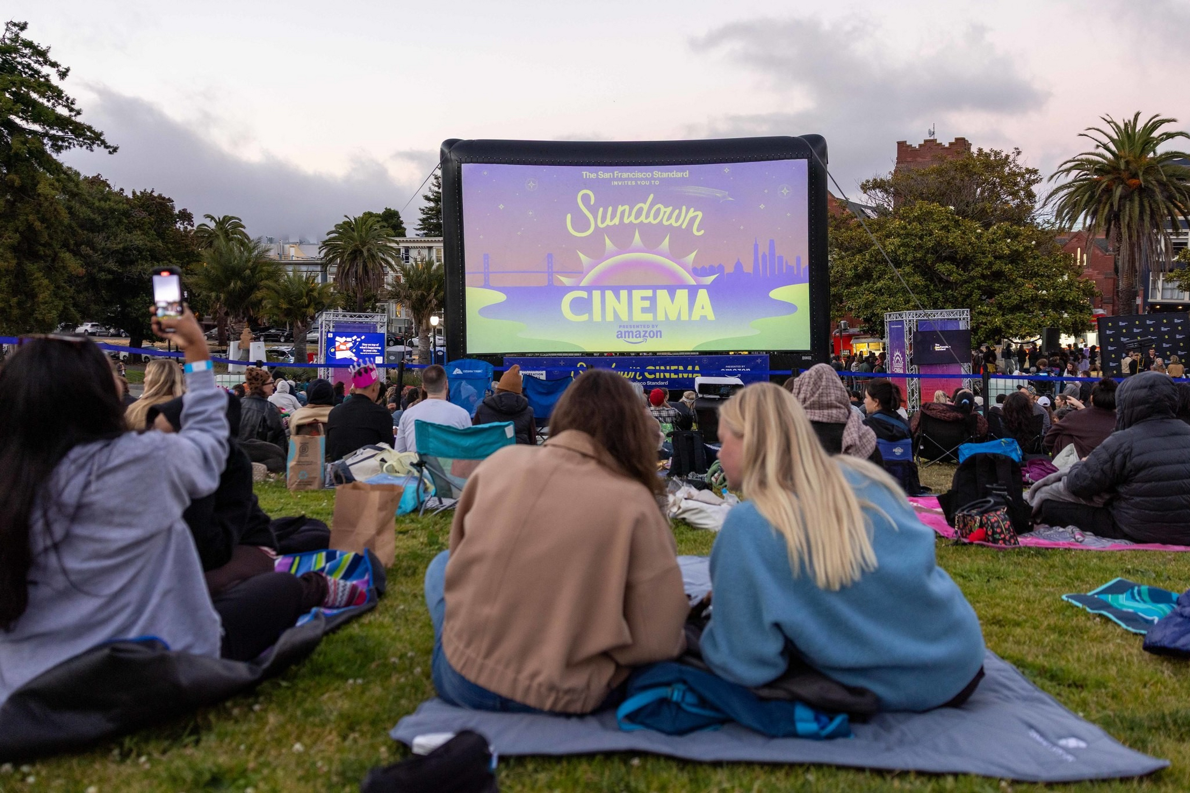 People sit on blankets on grass watching a large outdoor inflatable screen displaying “Sundown Cinema” in a park during early evening.