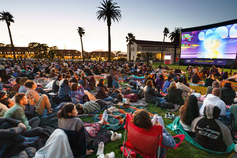 A large crowd sits on blankets and chairs on grass, watching an animated movie at an outdoor cinema during sunset, with palm trees and buildings in the background.