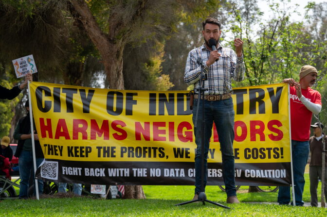 A man speaking into a microphone stands in front of a banner that reads "City of Industry harms neighbors. They keep the profits, we pay the costs" as others stand around.