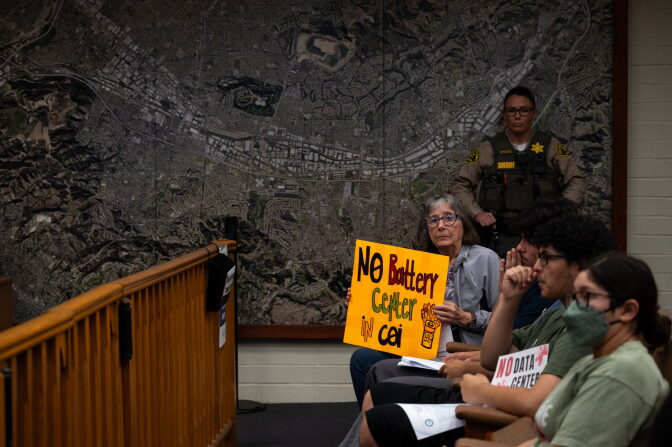 People sit in a room as one person holds a sign that reads "no battery center in Coi." A sheriff deputy stands in the background by the wall.