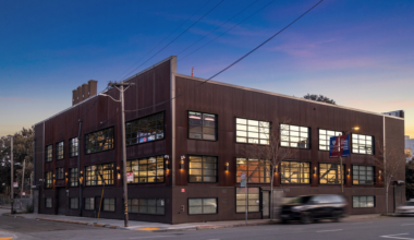A two-story brown industrial-style building sits on a street corner at dusk, with large windows and blurred cars passing by.