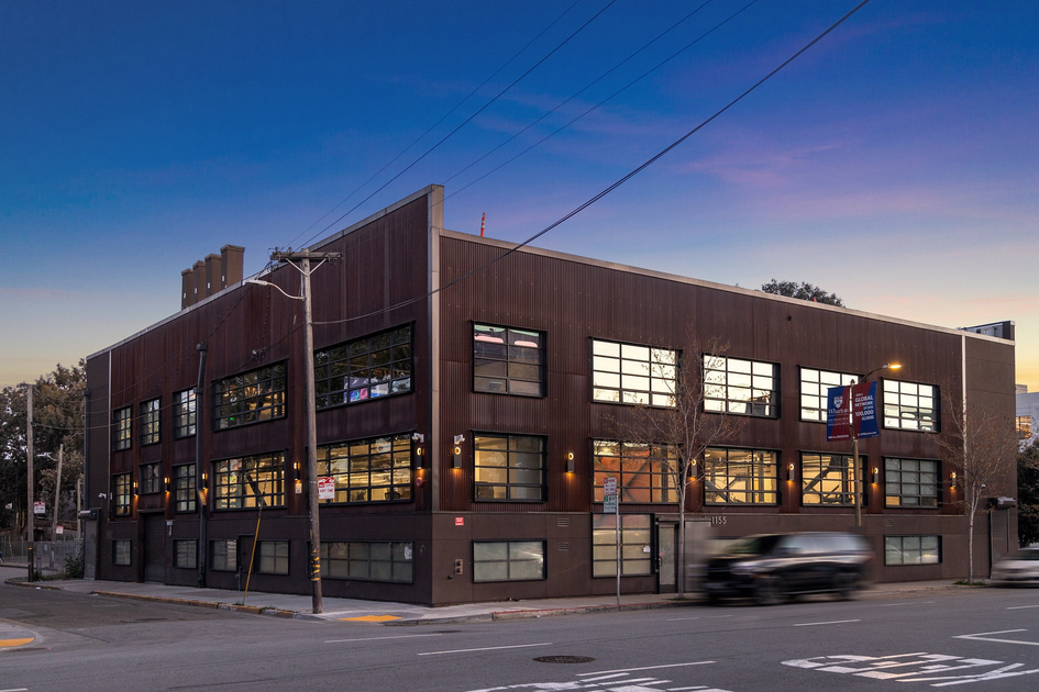 A two-story brown industrial-style building sits on a street corner at dusk, with large windows and blurred cars passing by.