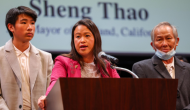 A woman in a pink jacket speaks at a podium, flanked by a young man in a light suit and an older man wearing a partially lowered face mask.