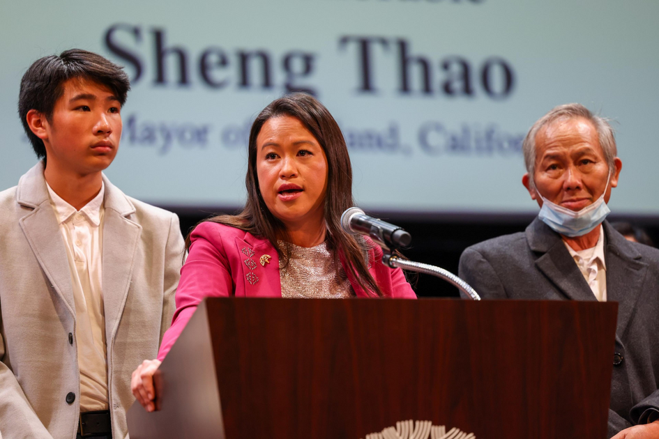 A woman in a pink jacket speaks at a podium, flanked by a young man in a light suit and an older man wearing a partially lowered face mask.