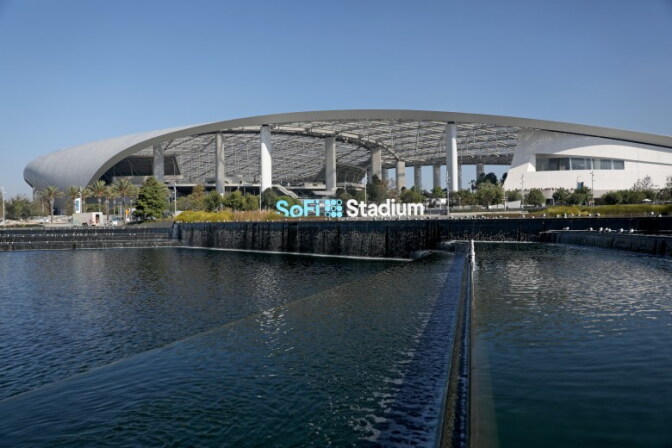 The exterior of SoFi Stadium stands in daylight under a cloudless blue sky. A wide, sweeping white roof canopy arches over the building, supported by tall white columns, with a translucent panel structure visible beneath it.