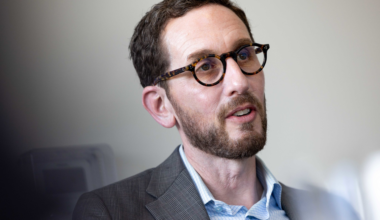 A man with short dark hair, a beard, round tortoiseshell glasses, and a plaid blazer over a light blue shirt is speaking or listening.