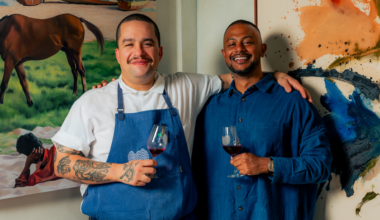 Two men smiling with arms around each other, holding glasses of red wine, one wearing a white shirt and blue apron, the other in a blue shirt.