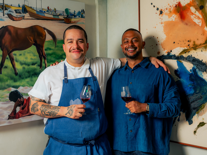 Two men smiling with arms around each other, holding glasses of red wine, one wearing a white shirt and blue apron, the other in a blue shirt.