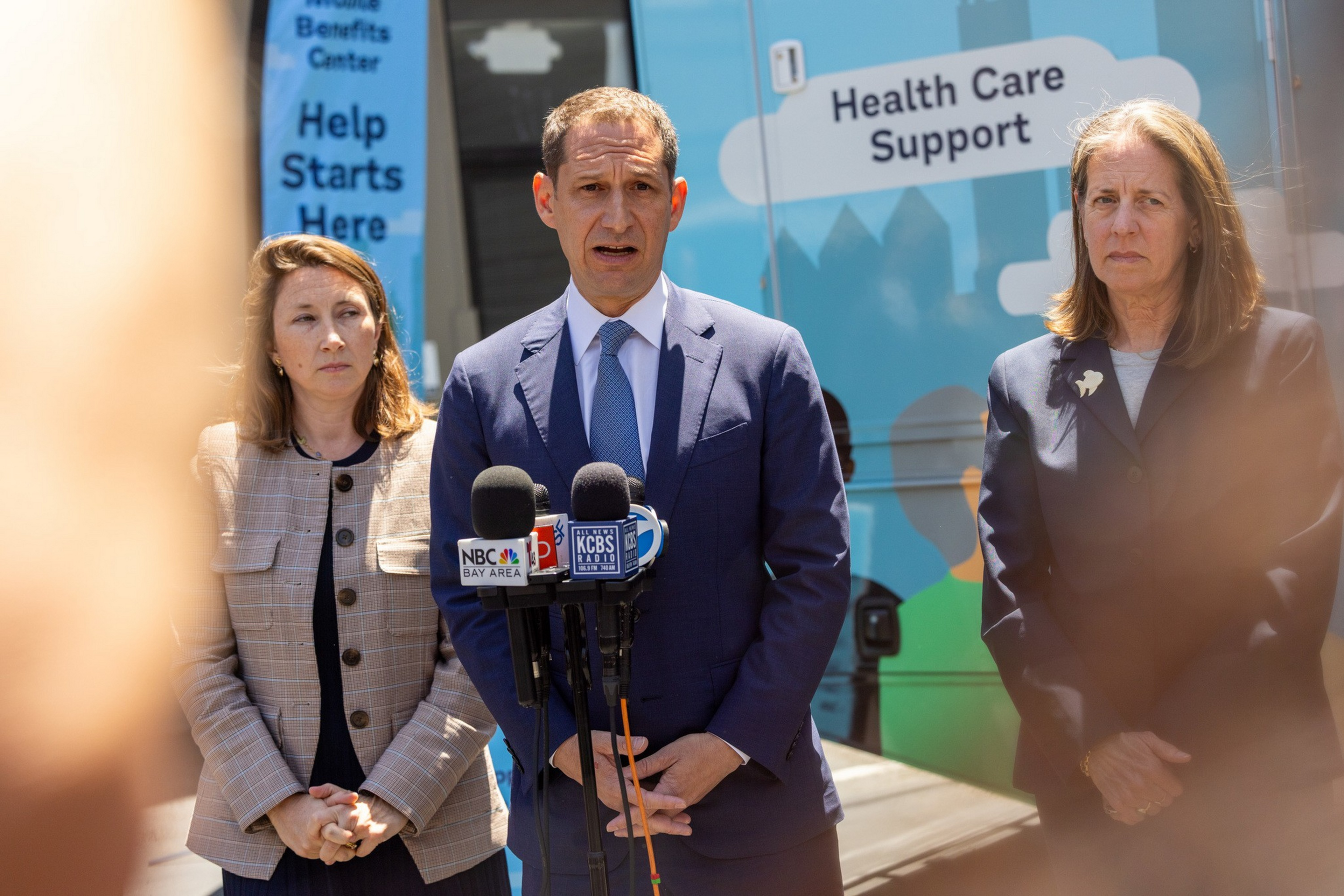 A man in a blue suit speaks into multiple microphones with two women standing on either side of him in front of a health care support vehicle.