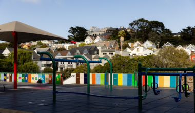 A colorful playground with climbing structures and swings sits empty in front of a row of residential houses under a clear sky.