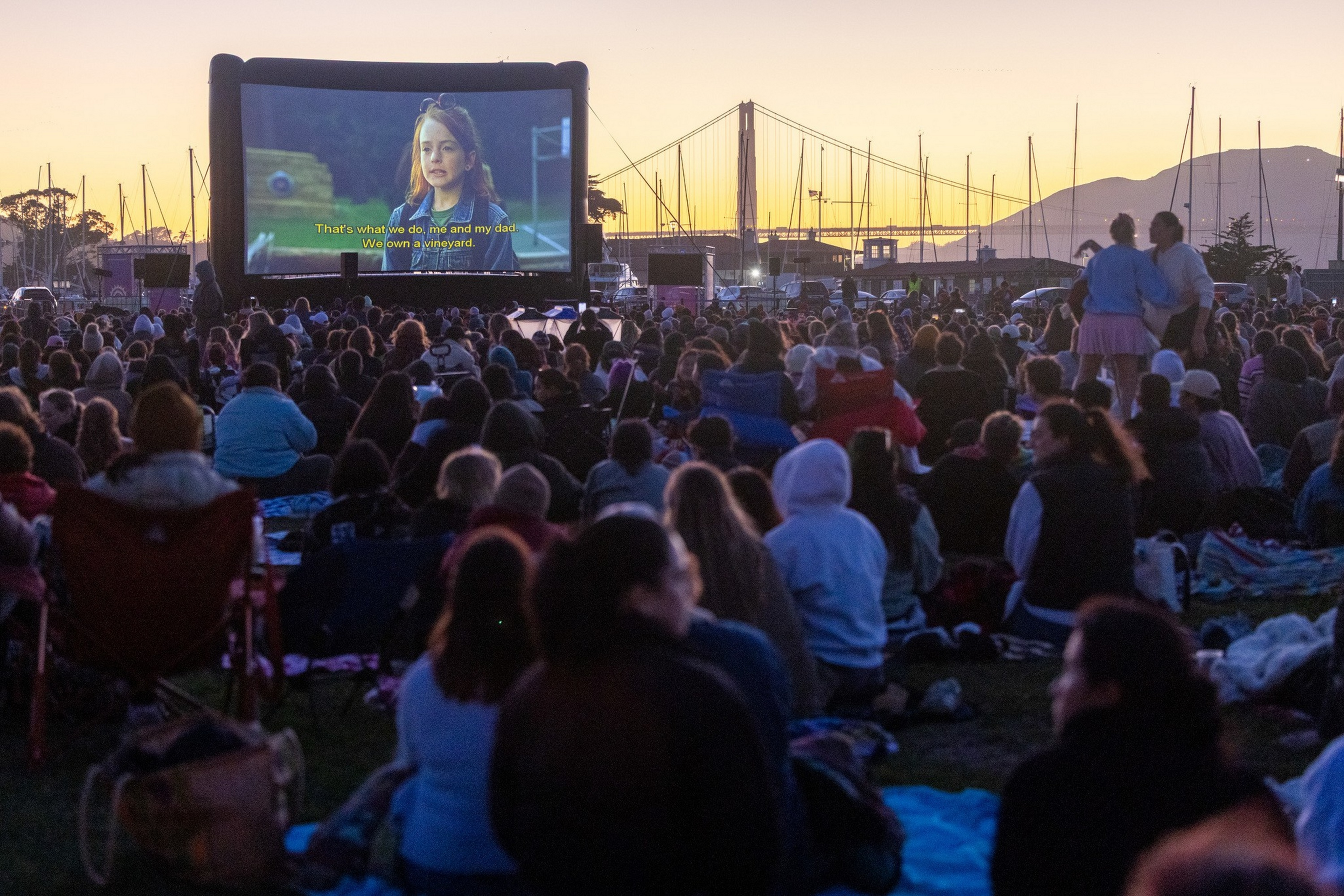 A large crowd sits on blankets and chairs outdoors, watching a movie on a big screen at sunset with a bridge and boats in the background.