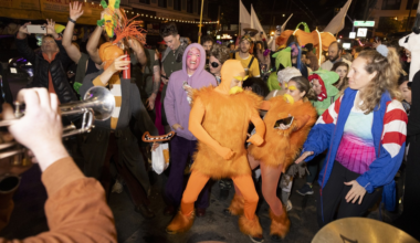 A large group of people and dogs, some in costumes, gather outdoors in front of a double-decker bus with Halloween decorations, including a large spider.