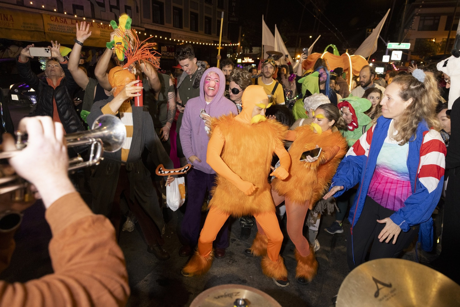 A large group of people and dogs, some in costumes, gather outdoors in front of a double-decker bus with Halloween decorations, including a large spider.