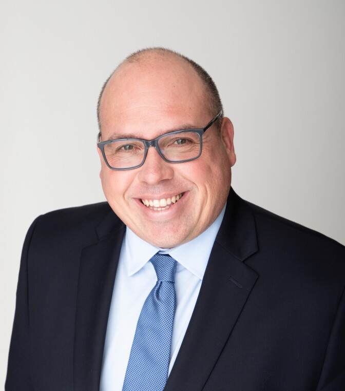 A person wearing glasses and a dark suit with a blue tie smiles for a headshot against a light gray background.