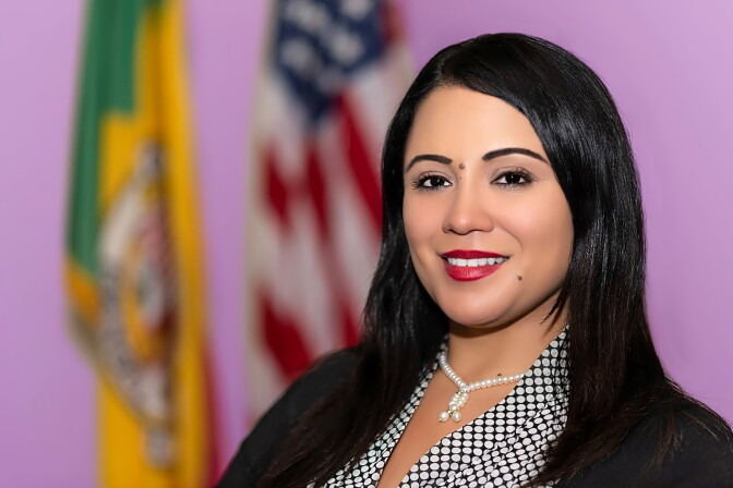 A person with long dark hair and red lipstick smiles, wearing a pearl necklace and polka-dot top, with the flags of the U.S. and Los Angeles behind her.