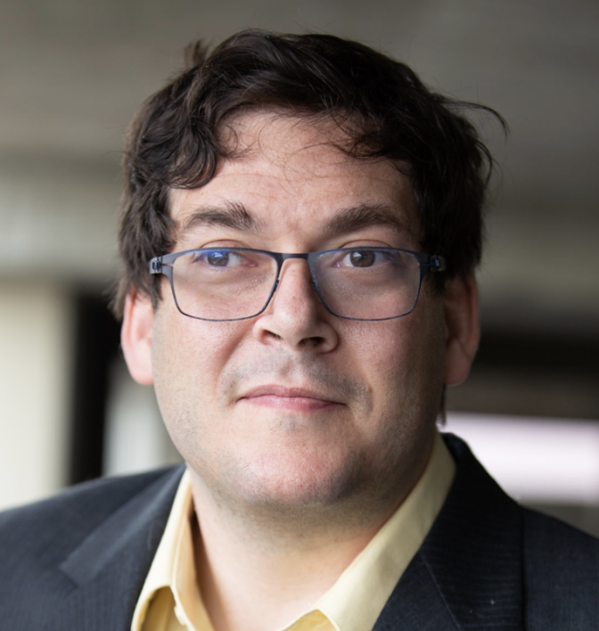 A person with curly dark hair and glasses wears a yellow shirt and dark blazer, posing for a close-up portrait.