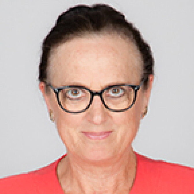A person with short dark hair and glasses wears a coral-colored top, smiling slightly in a studio portrait.