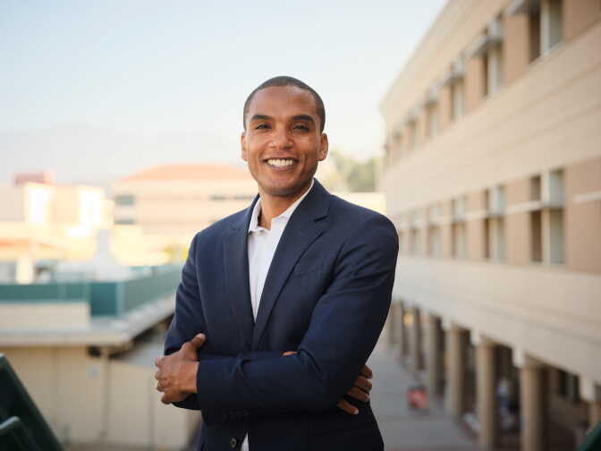 A person in a navy blazer smiles with arms crossed on an outdoor walkway between buildings.