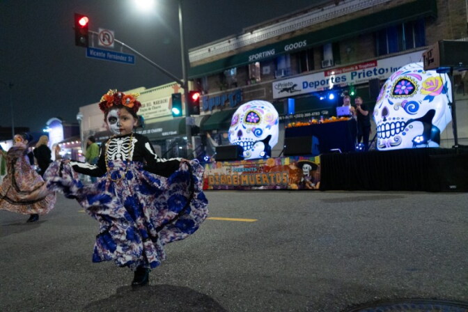 A child dances folkorico a party taking place at a closed off street intersection in front of a stage and businesses.