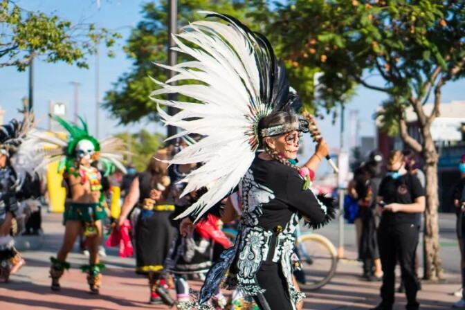 Dancers perform Danza Azteca outside, with one person wearing a white and black-colored outfit.