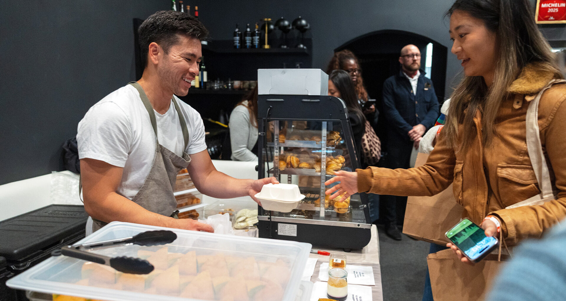 Asian man in a chef's apron hands baked goods to a customer.