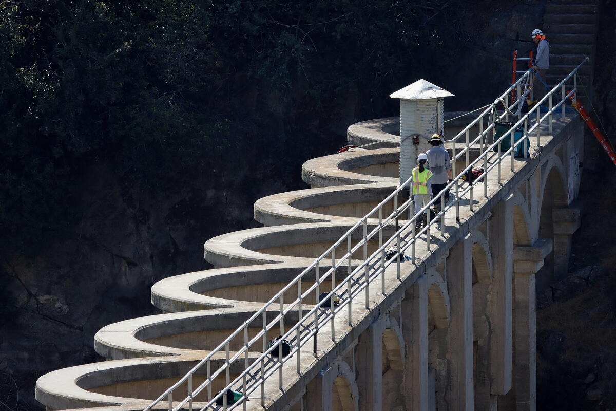 Three workers wearing hard hats stand along a narrow walkway with metal railings atop a concrete dam. 