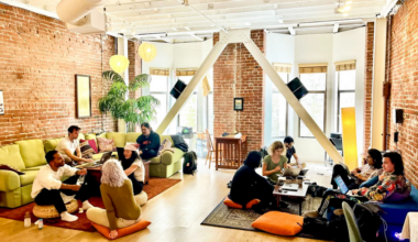 People work and chat in a bright, spacious room with exposed brick walls, green couches, cushions on the floor, plants, and laptops on low tables.