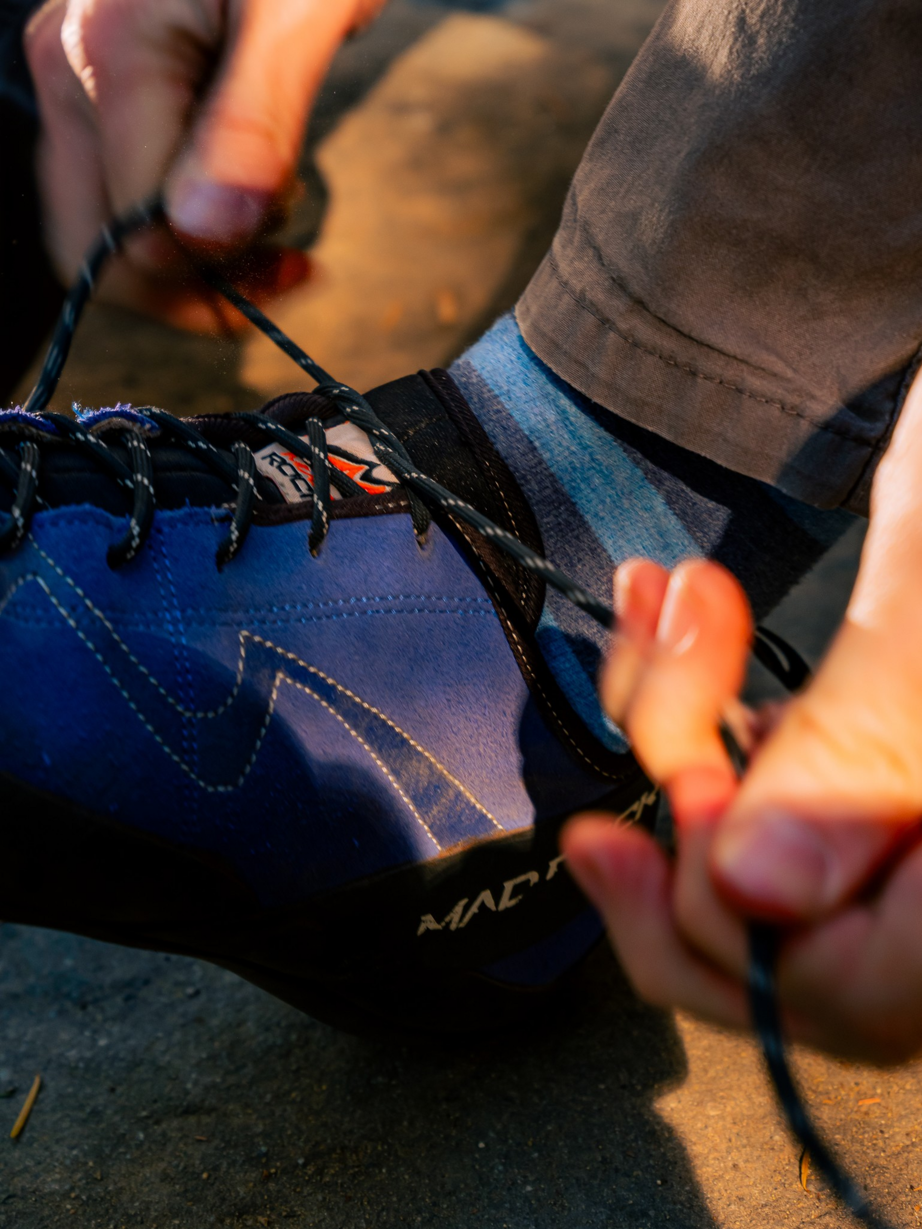 A person is tying the black laces of a blue and black climbing shoe while wearing striped socks and brown pants.