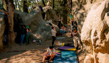Several people are bouldering and resting among large rocks in a wooded area with climbing mats spread on the ground for safety.