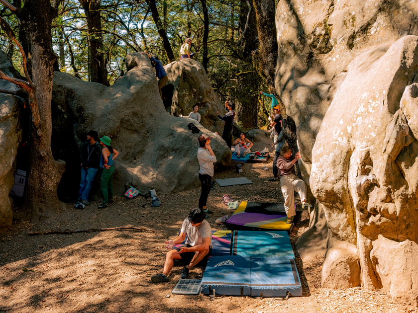 Several people are bouldering and resting among large rocks in a wooded area with climbing mats spread on the ground for safety.