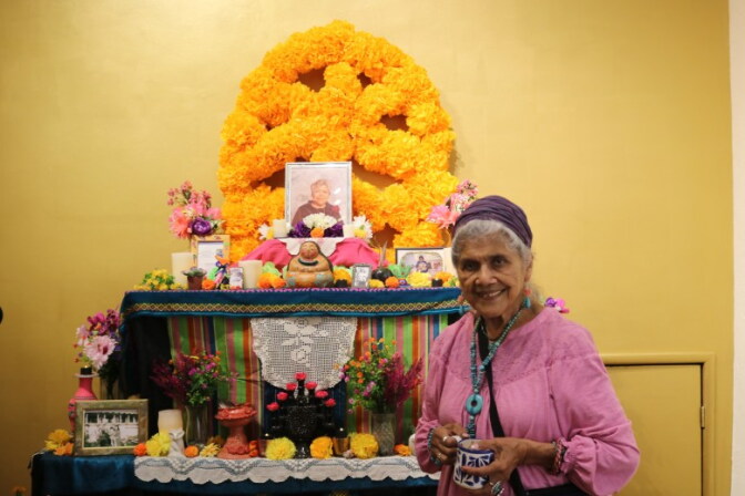 Ofelia Esparza, an older woman with medium skin tone, wearing a pink shirt and jewelry, stands by an altar with photos and flowers.