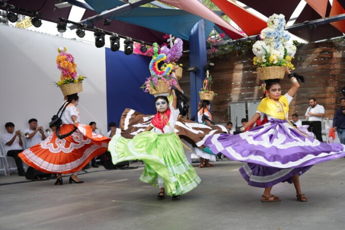 Young dancers wearing colorful dress and face paint dance as they hold a flower arrangement on their heads.