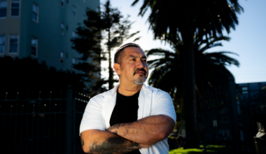 A man with tattoos and a short beard stands with arms crossed, wearing a white short-sleeve shirt over a black t-shirt, with palm trees and buildings behind him.