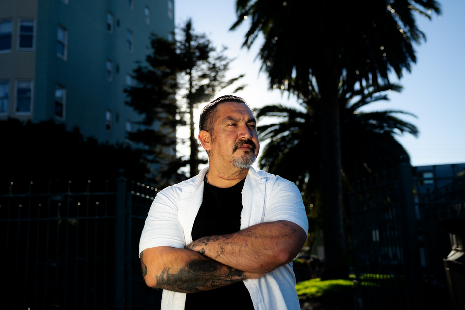 A man with tattoos and a short beard stands with arms crossed, wearing a white short-sleeve shirt over a black t-shirt, with palm trees and buildings behind him.
