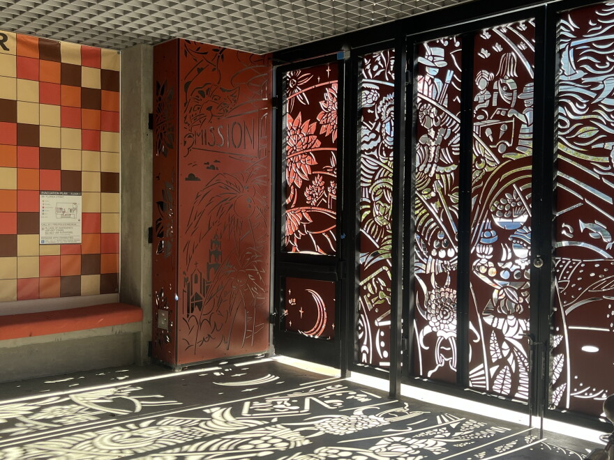 The intricate rust-red front gate of Cultura y Arte Nativas de las Americas, or CANA, casts shadows in the shapes of kids, plants, bird, and the moon on the ground.