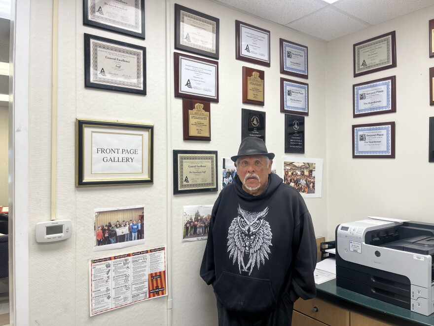 Journalist Juan Gonzales, a Latino man with a neat white beard, wears a crisp black fedora and a black hoodie with an owl on it. He stands against a wall of awards and accolades for City College's newspaper, The Guardian, in The Guardian's newsroom.