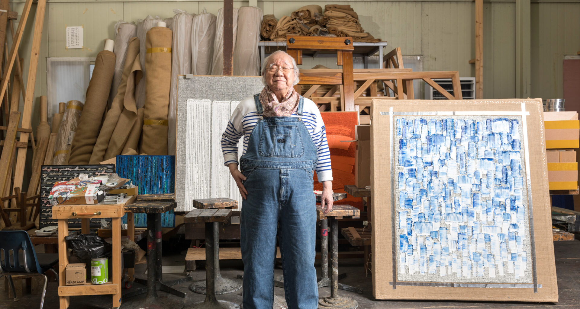 older Korean man in denim overalls next to abstract painting