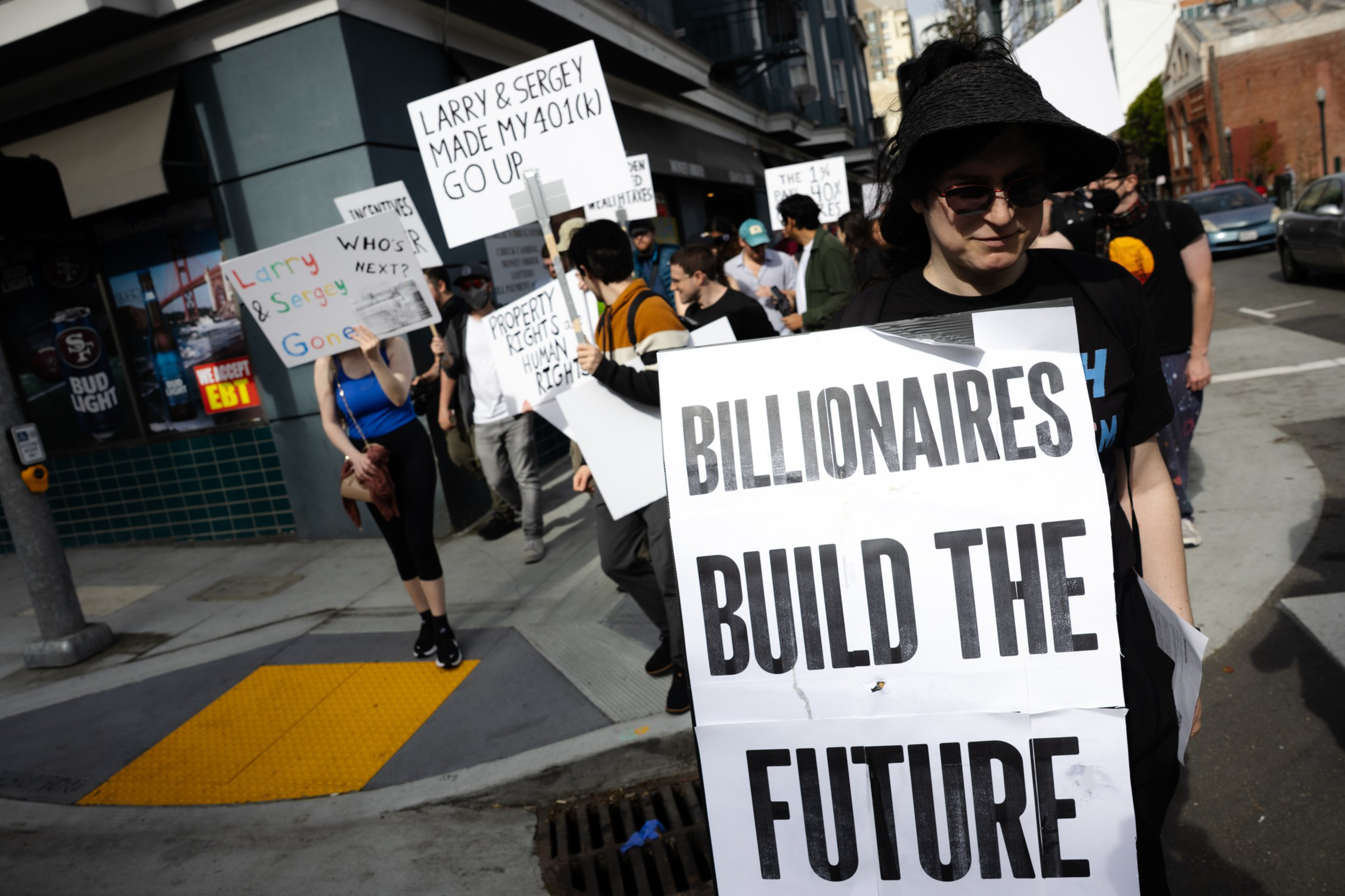 People march in a street protest holding signs with messages like “BILLIONAIRES BUILD THE FUTURE” and “LARRY & SERGEY MADE MY 401(K) GO UP.”