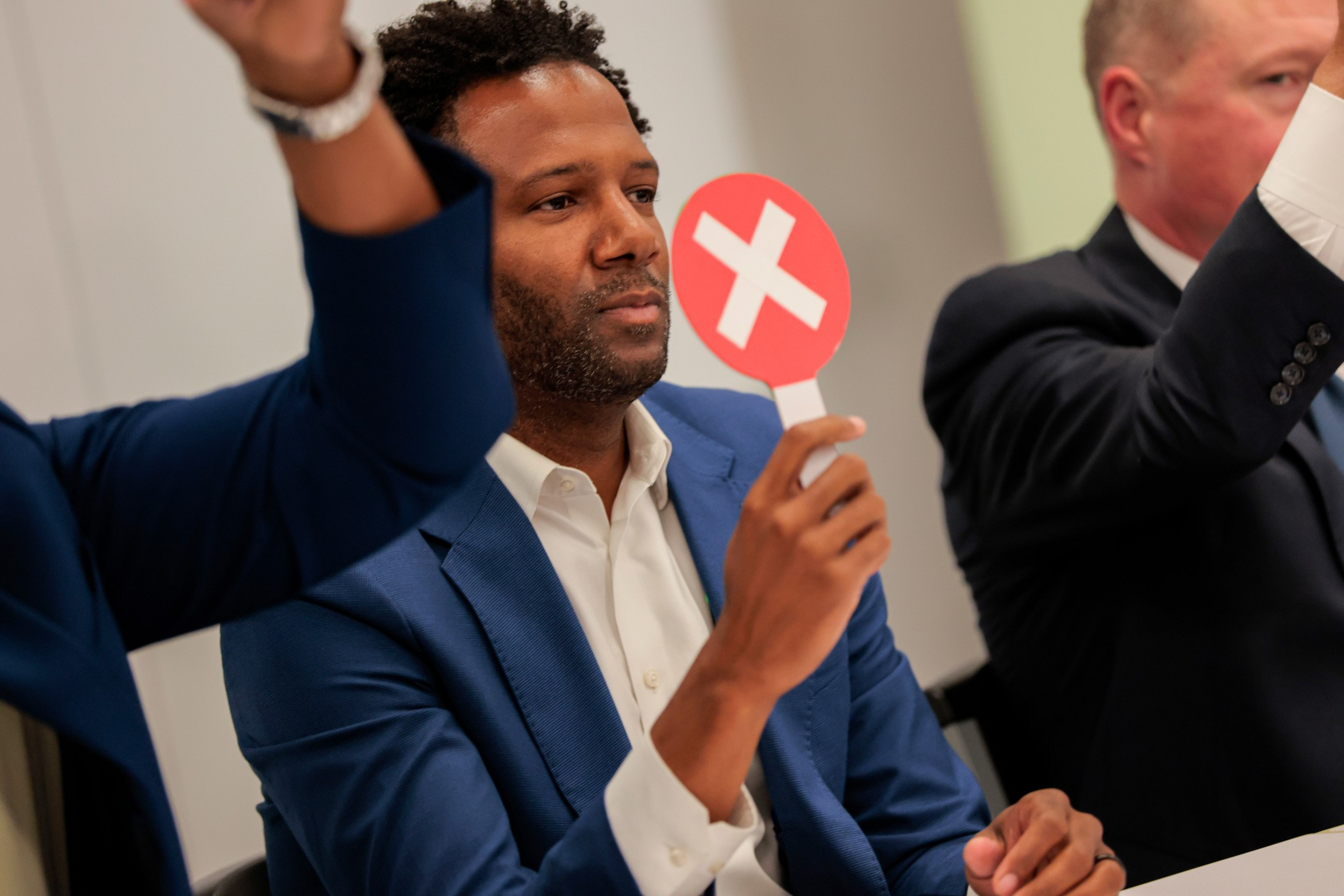A man in a blue suit holds up a red paddle with a white X, while others raise hands, suggesting voting or signaling disapproval.
