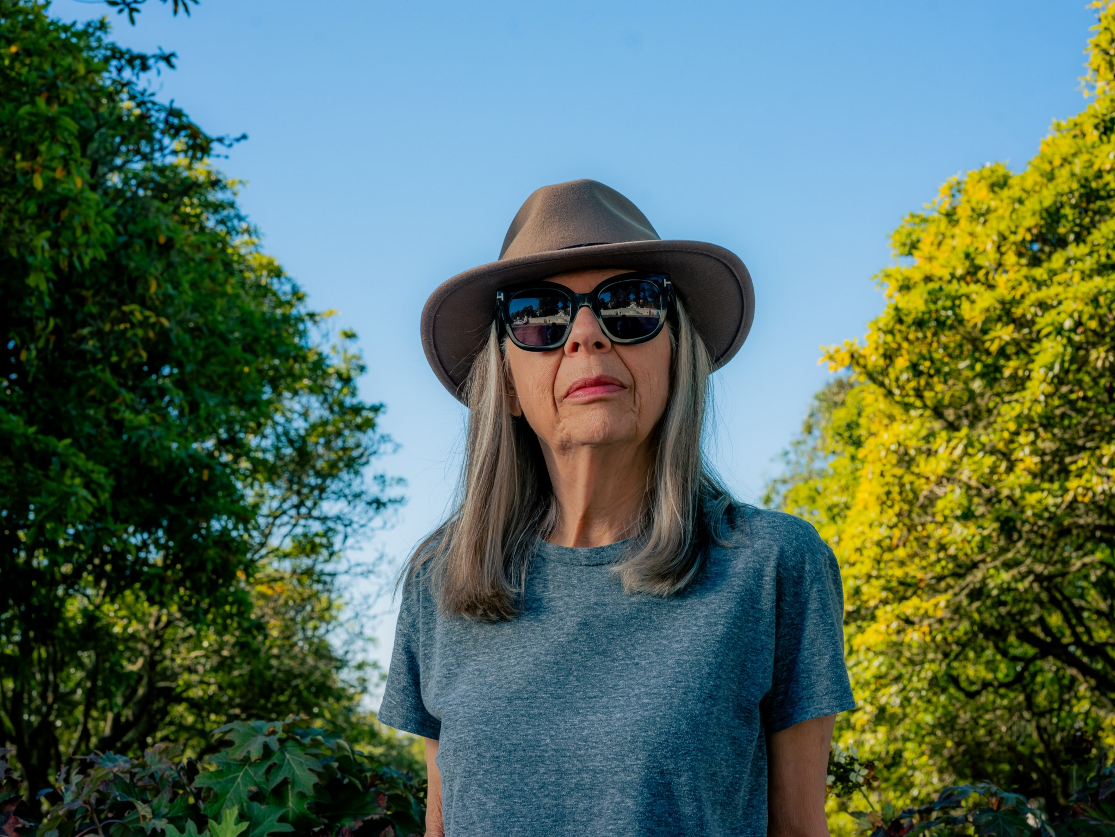 A woman with long gray hair wears a brown wide-brimmed hat, black sunglasses, and a gray t-shirt, standing outdoors with green trees around her.