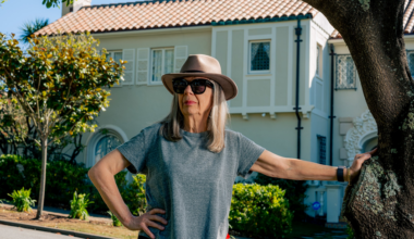 A woman wearing a gray t-shirt, sunglasses, and a beige hat stands by a tree, with one hand on her hip and a house with a tiled roof in the background.