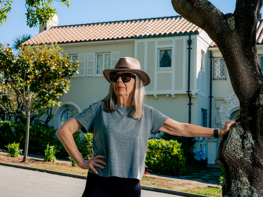 A woman wearing a gray t-shirt, sunglasses, and a beige hat stands by a tree, with one hand on her hip and a house with a tiled roof in the background.